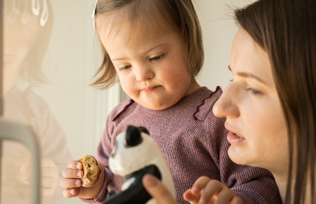 Portrait view of girl with down syndrome holding cookie and looking at her panda toy while standing near her mother.